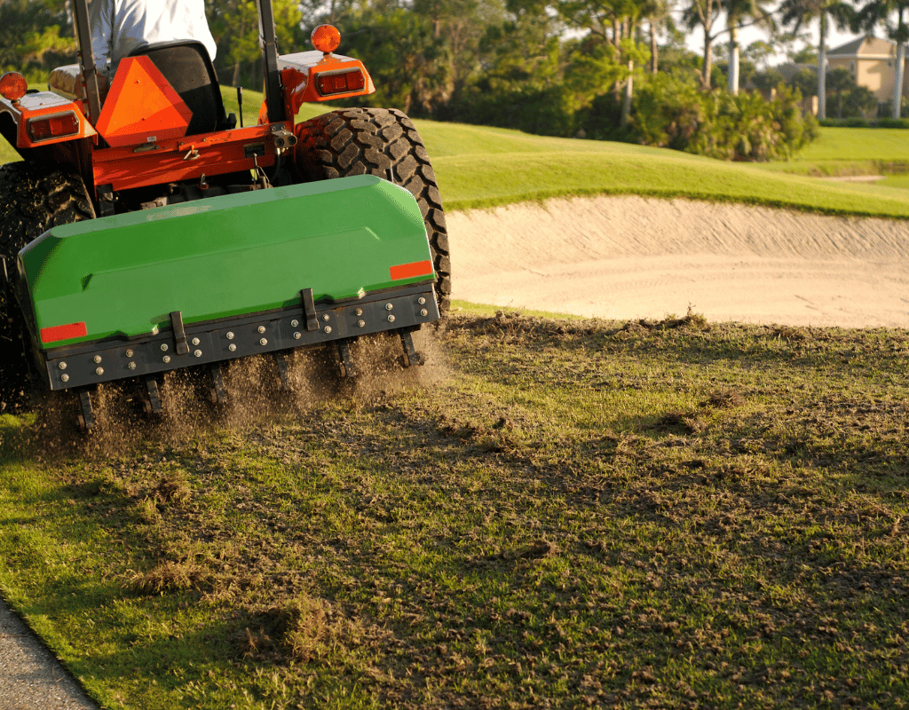Riding aerator machine punching cores into turf next to a sand bunker on a golf-style lawn