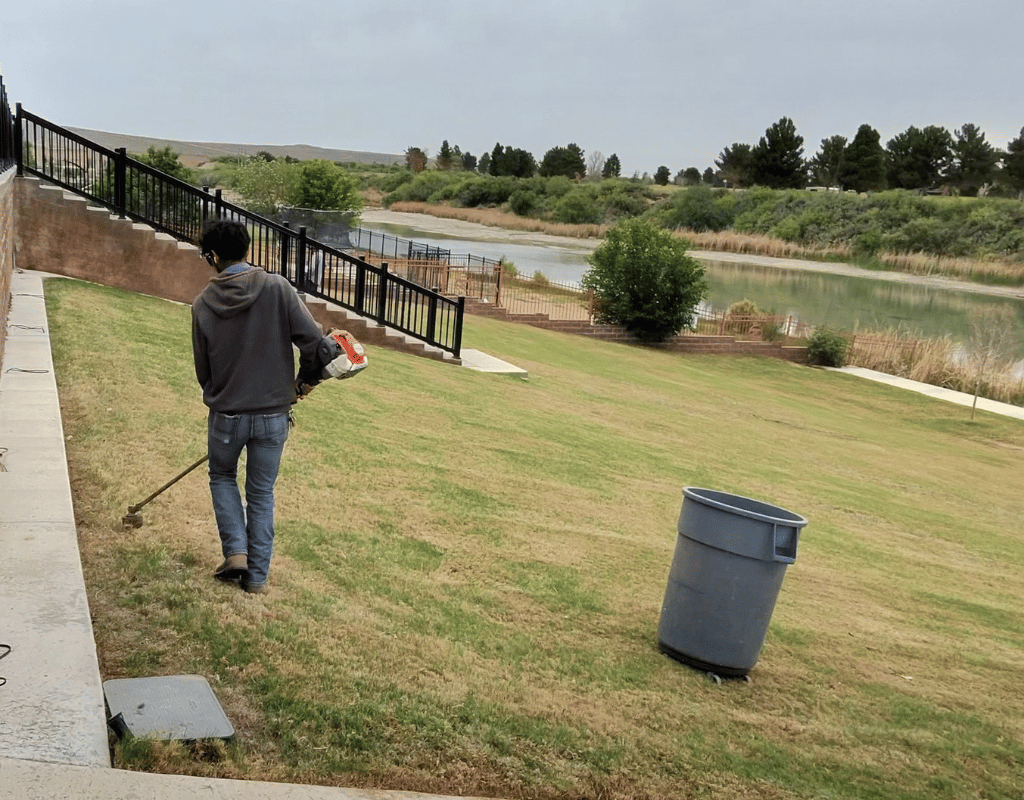 Landscaper using a string trimmer along a grassy area beside a retention pond and trash bin