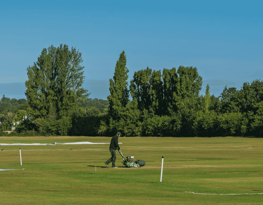 Groundskeeper pushing a mower across a wide open lawn bordered by tall trees on a commercial property