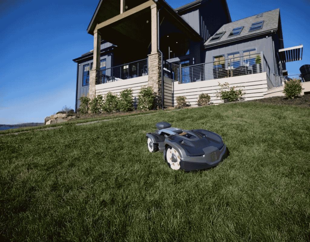 Black robotic lawn mower cutting grass on a sloped backyard lawn near a modern home